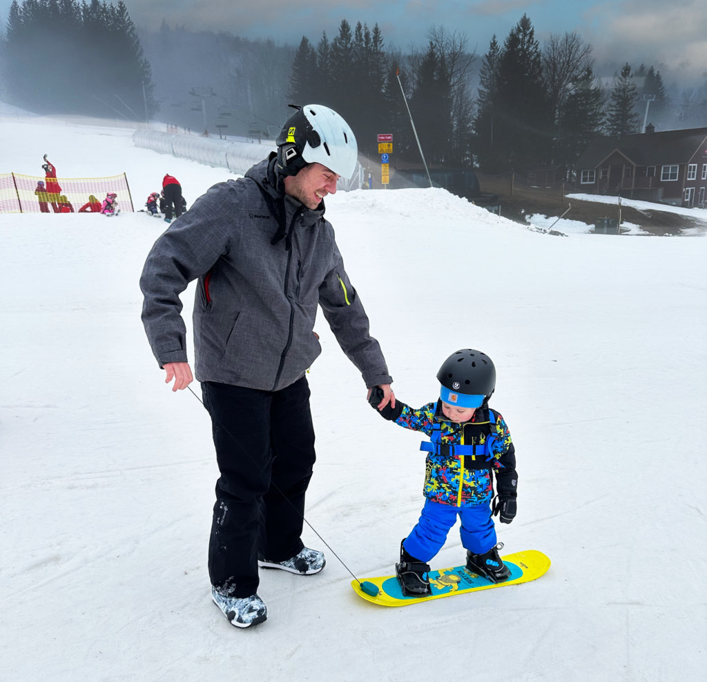 Father guiding toddler son on snowboard at Stratton